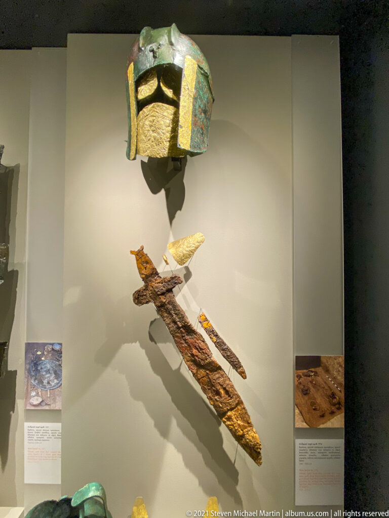 Green bronze helmet with gold trim above a long corroded iron weapon retaining bright gold fittings.