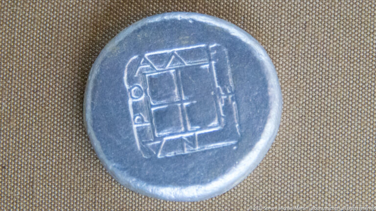 Close-up of a dark ancient coin reverse with a rectangular geometric design, angular lines, and a small circular element.