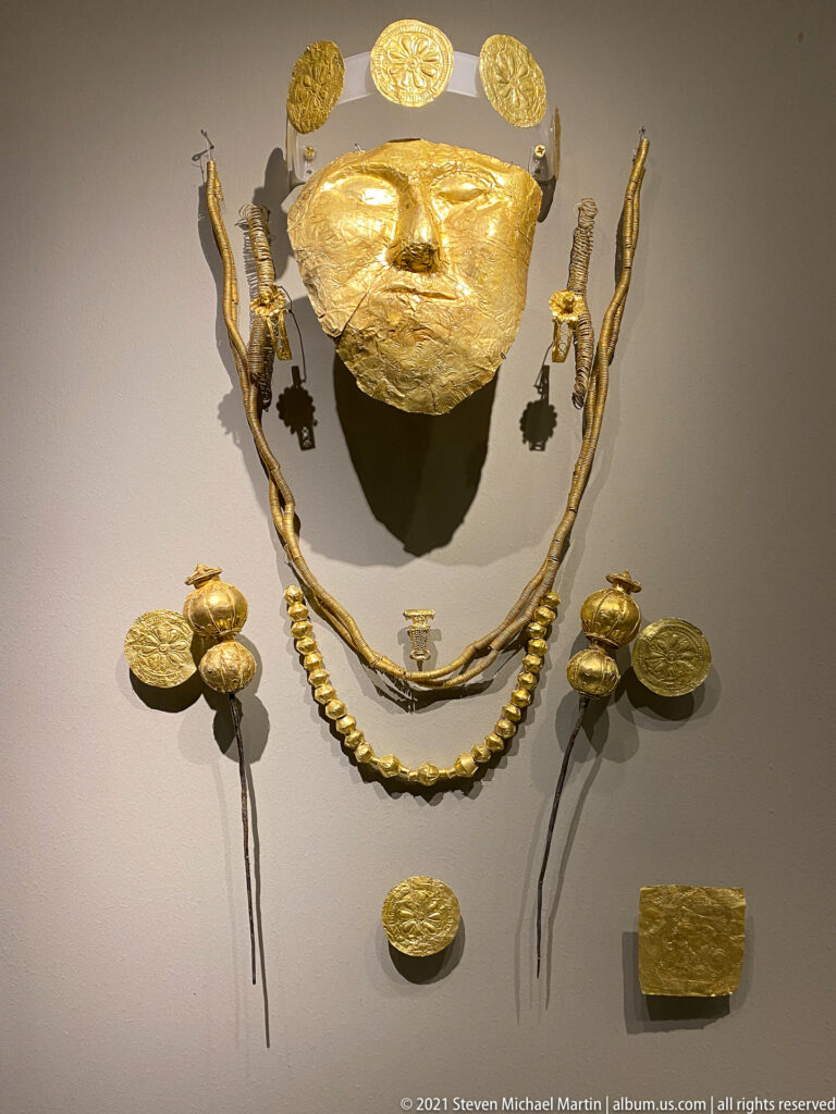 Museum display of a hammered gold face covering with necklaces, pendants, circular appliqués, and other gold funerary ornaments.