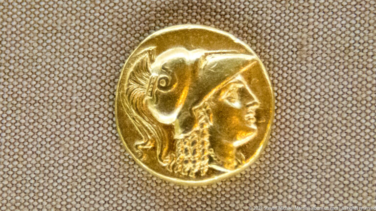 Round gold coin with a helmeted profile in relief against a textured museum display.