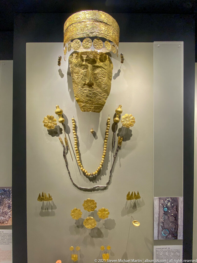 Elaborate gold burial display with hammered face covering, broad headband, layered necklaces, rosettes, disks, and triangular ornaments.