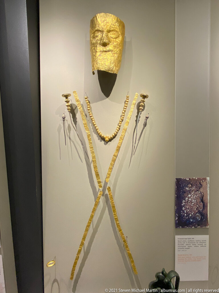 Gold face covering above two necklaces, crossed gold bands, pins, and small ornaments arranged vertically in a museum case.