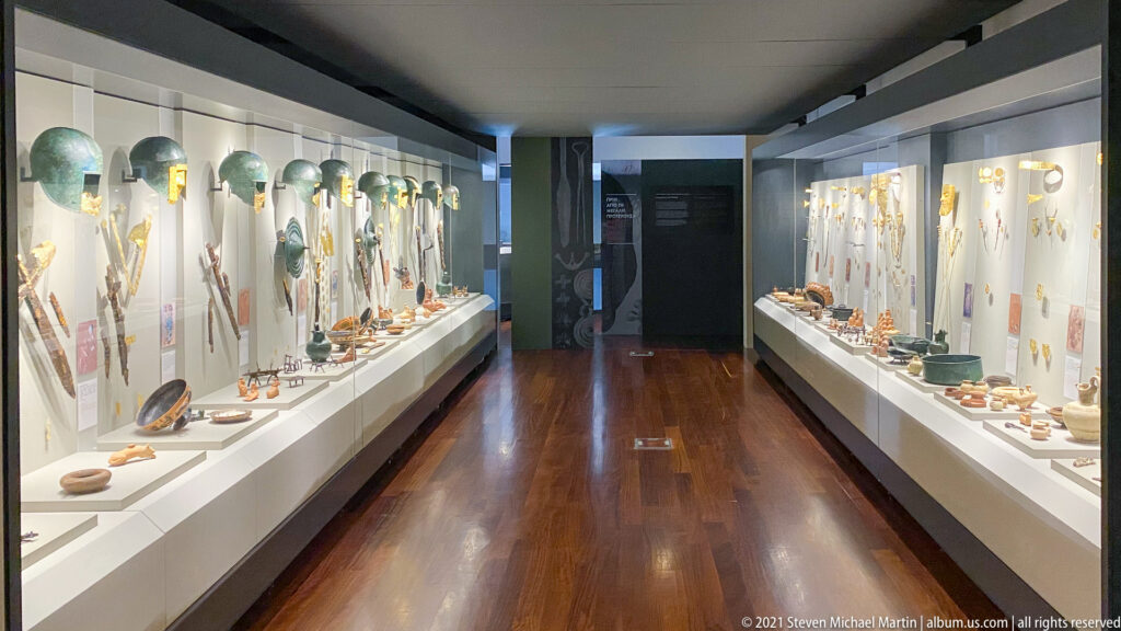 Long museum gallery with illuminated display cases of pottery, metalwork, jewelry, and archaeological finds on both sides.