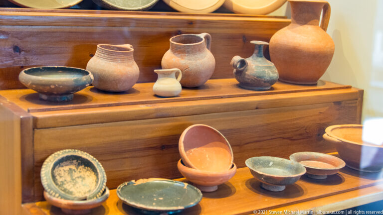 Ancient bowls, cups, jugs, and jars of varied shapes arranged on stepped museum shelves.