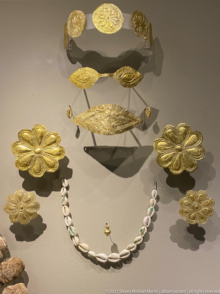 Gold rosettes and patterned bands displayed above a necklace of pale shell-shaped beads.