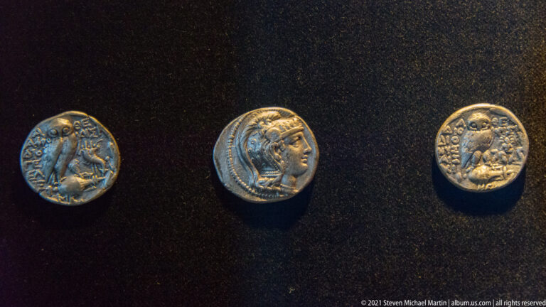 Three ancient silver coins with profile heads and inscriptions displayed against black fabric.