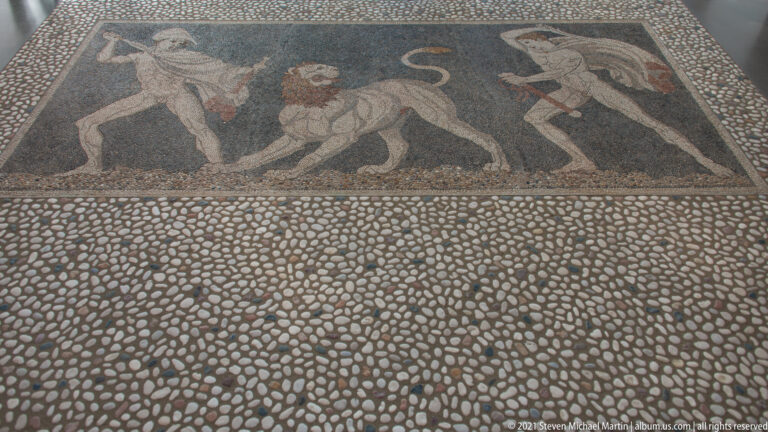 Rectangular figural pebble mosaic bordered by a broad field of densely packed pale and dark stones.