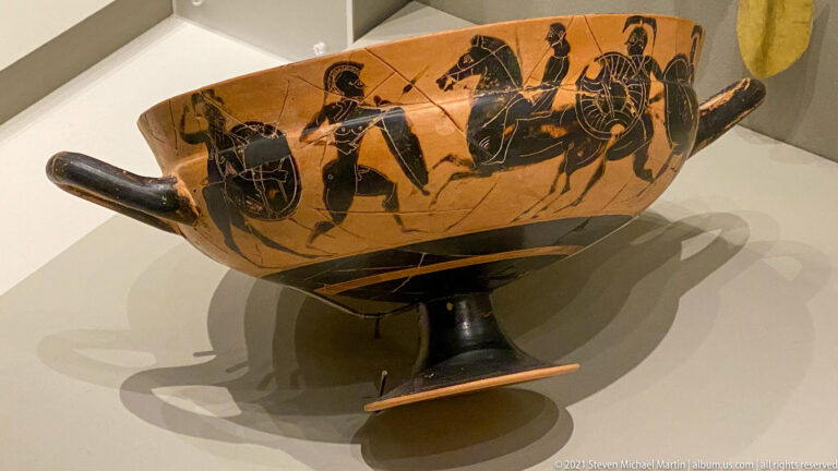Orange-and-black stemmed drinking cup with human figures, horses, and a wheeled vehicle painted around the bowl.