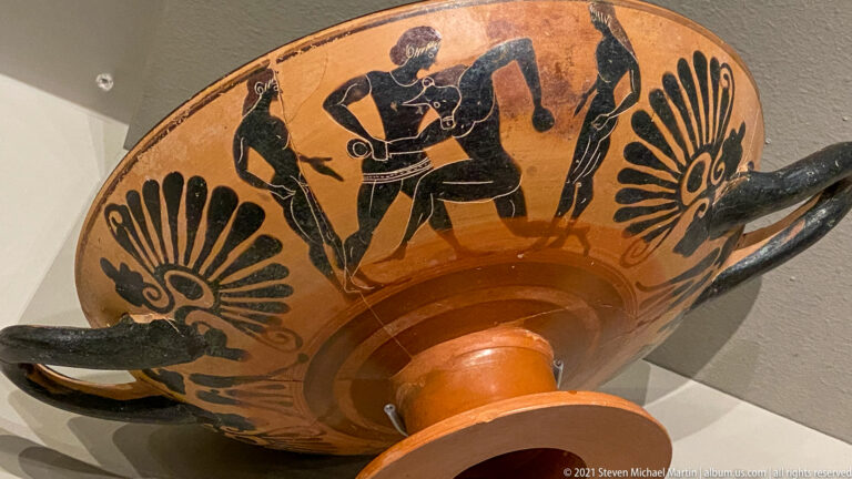 Black-figure stemmed cup seen at an angle, with human figures and large palmettes between horizontal handles.