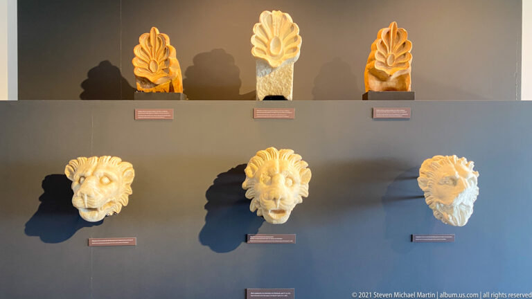Three upright palmette ornaments above three projecting lion-head architectural terracottas on a museum wall.