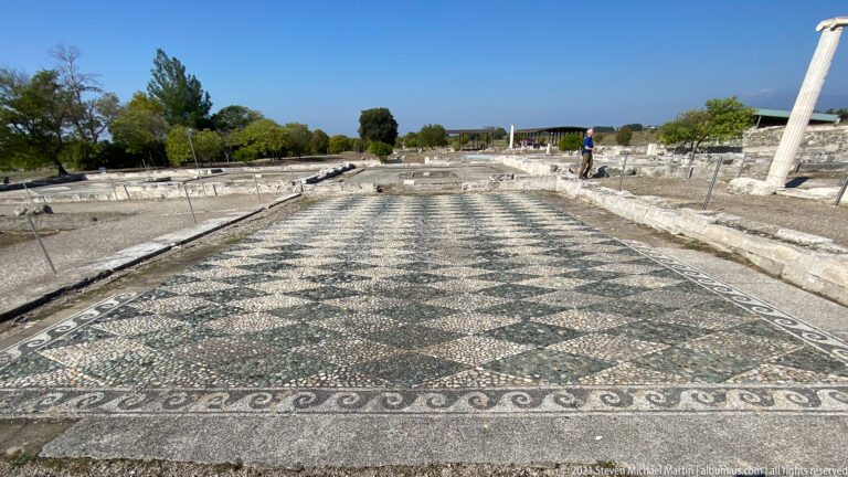 Long in-situ pebble mosaic floor with a repeating geometric lattice and decorative borders amid excavated walls.