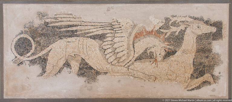Winged four-legged hybrid creature with a long curling tail rendered in pale, dark, and reddish pebbles.