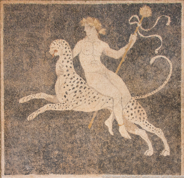 Human figure riding a spotted feline and holding a long staff in a dark-ground pebble mosaic.