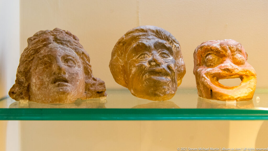 Three small sculpted theatrical heads with strongly contrasting expressions arranged on a glass shelf.