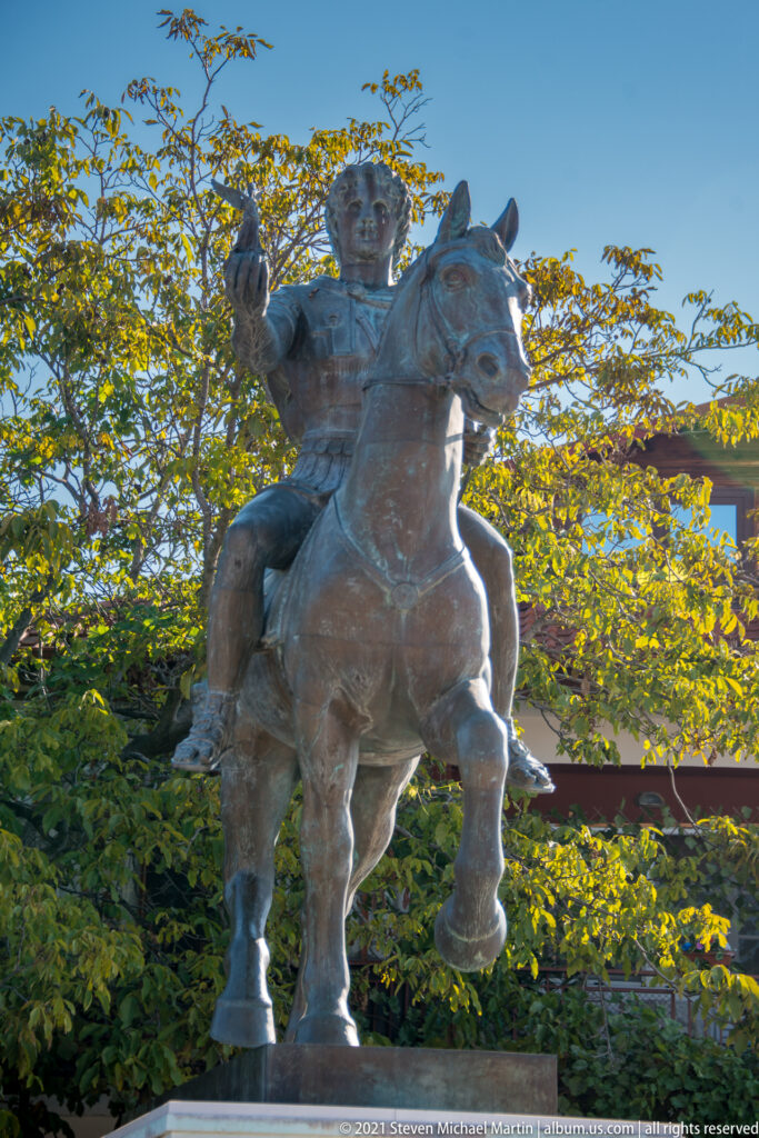 Backlit bronze equestrian statue of a gesturing male rider on a stepping horse, framed by green trees and blue sky.