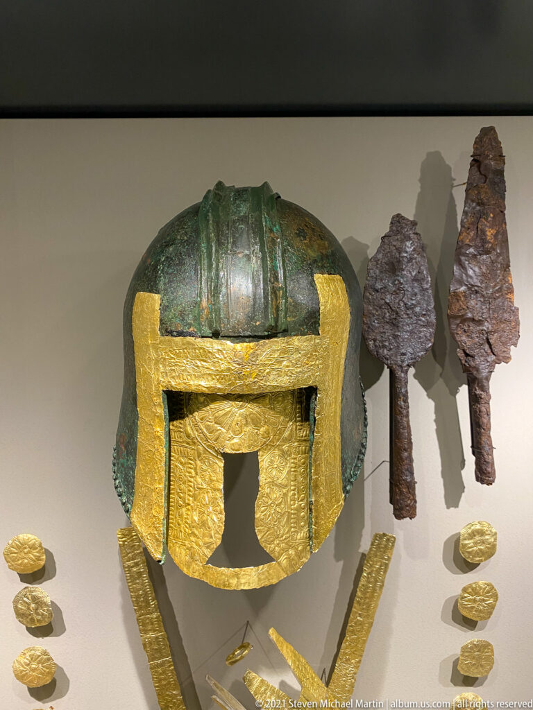 Green-patinated bronze helmet with gold facing, iron weapon heads, gold bands, and small gold disks in a museum display.