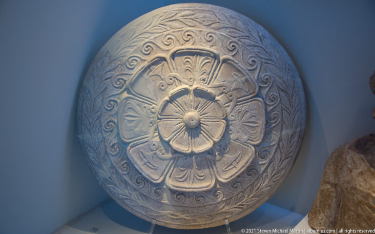 Round gray relief object with a central rosette, radiating palmettes, and scrolling outer border.