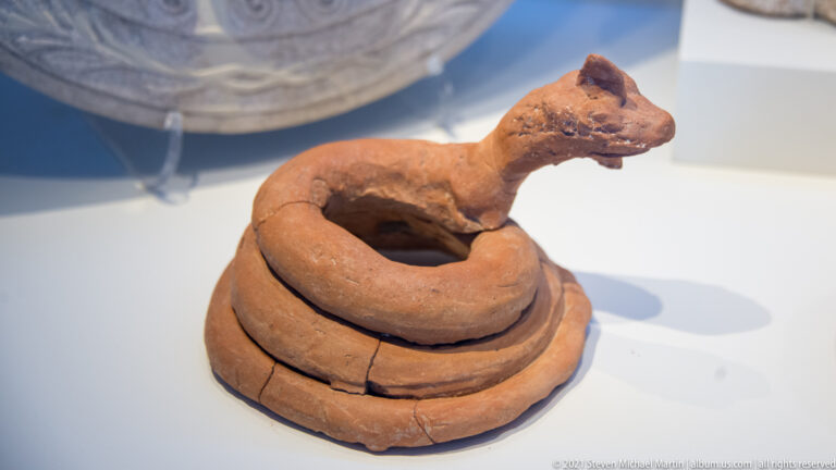 Reddish terracotta object formed as a coil with a long-necked animal head rising from one side.