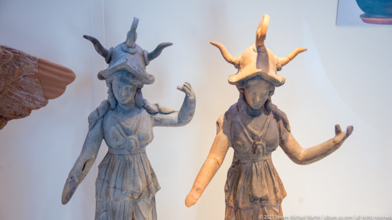 Two standing terracotta female figurines wearing tall headgear and belted garments, each posed with raised arms.