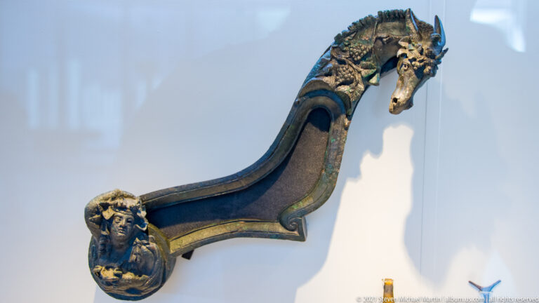 Curved dark metal fitting with an ornate animal head at one end and a small face at the other.