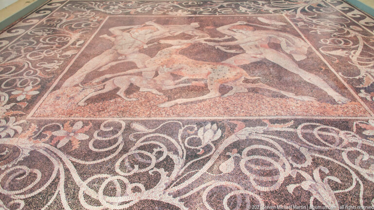 Rectangular pebble mosaic with a central hunting scene framed by broad scrolling vegetal borders.