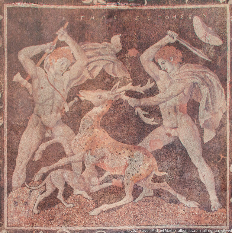 Close view of a pebble mosaic showing two hunters surrounding a spotted animal, with an inscription along the top.