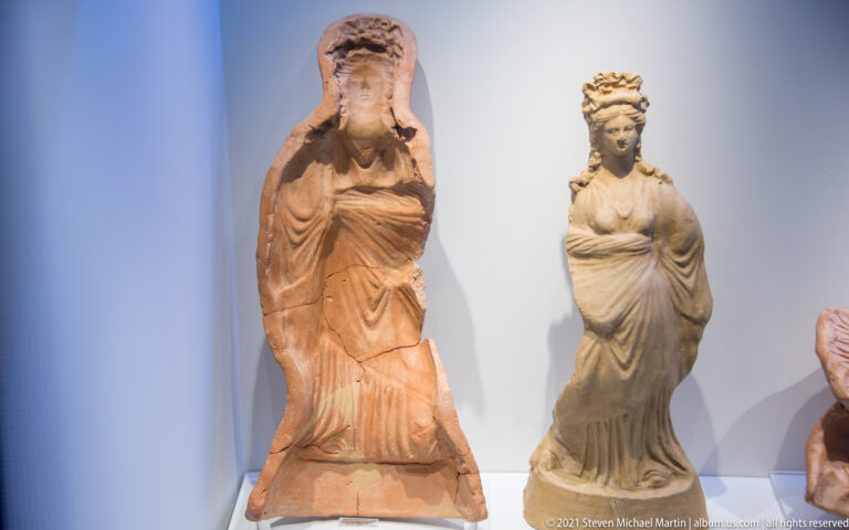 Two standing terracotta female figurines in long draped garments and elaborate headdresses.
