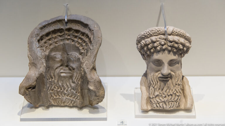 Two bearded male sculpted heads, one with a hood-like frame and one wearing textured rounded headgear.