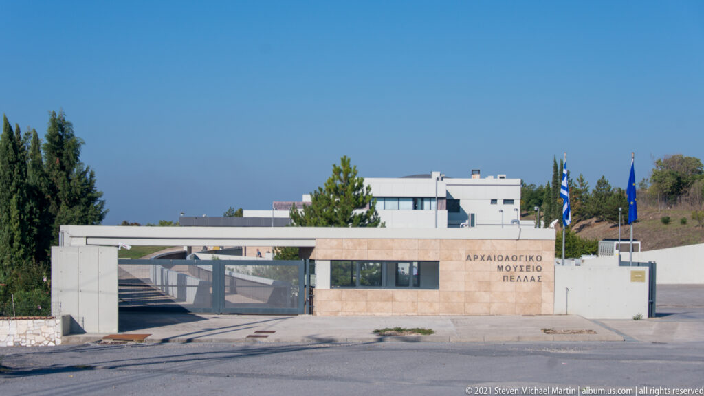 Low modern entrance of the Archaeological Museum of Pella with Greek and EU flags and Greek lettering on the stone façade.