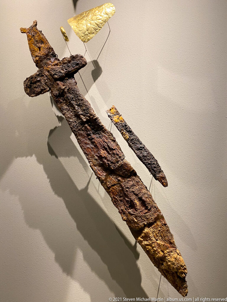 Corroded iron blade and weapon fragments mounted diagonally with surviving gold fittings in a museum case.