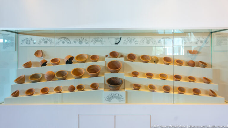 Dozens of small ancient ceramic bowls and cups arranged in rows on stepped museum shelves.
