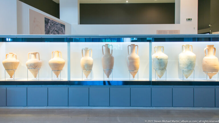 Row of tall amphorae with paired handles displayed upright in a long museum case.