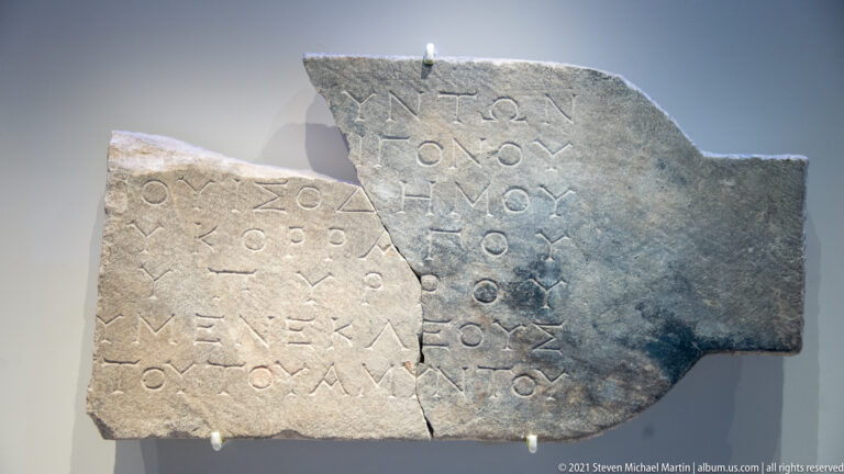 Fragmentary gray stone slab with multiple lines of carved Greek text and a vertical repaired break.