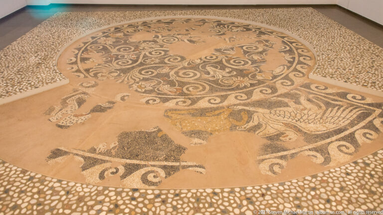 Circular pebble mosaic with a spiral-patterned center and surrounding human and animal figures.