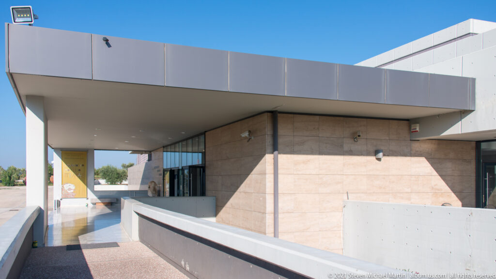 Covered concrete ramp leading to glass doors beneath a broad cantilevered canopy at the Archaeological Museum of Pella.