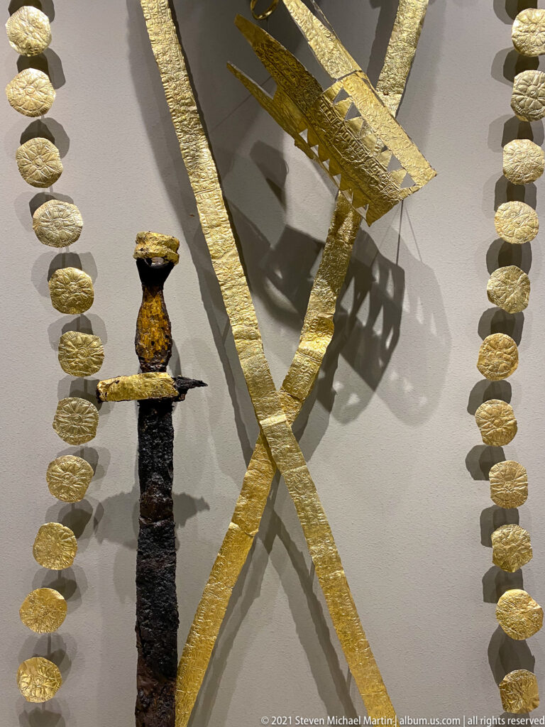 Corroded vertical iron weapon flanked by rows of gold disks and crossed gold-covered bands in a museum display.