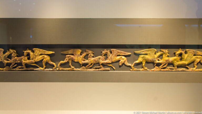 Narrow gilded relief frieze with horsemen, fallen warriors, and overlapping combatants on a dark ground.