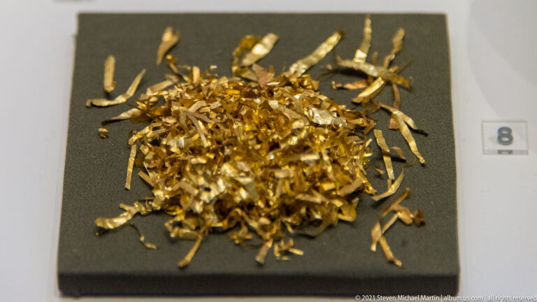 Pile of thin crumpled gold strips and cut fragments scattered across a dark museum display.