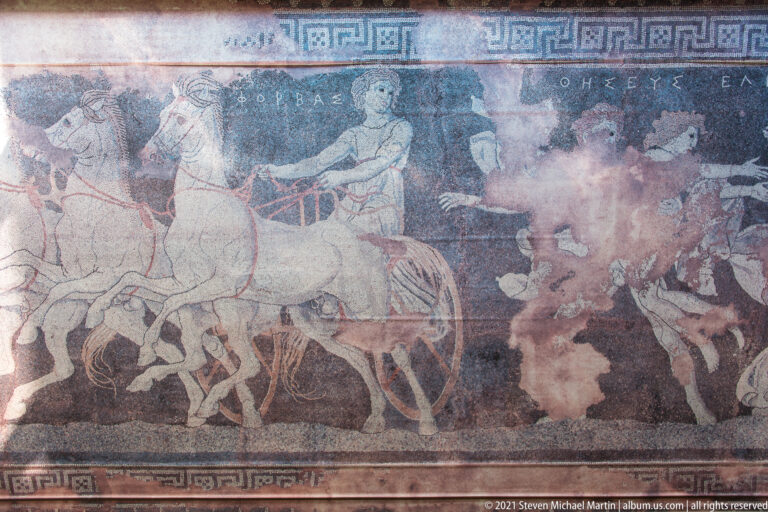 Fragmentary painted panel with a multi-horse chariot scene, human figures, inscription, and geometric border.