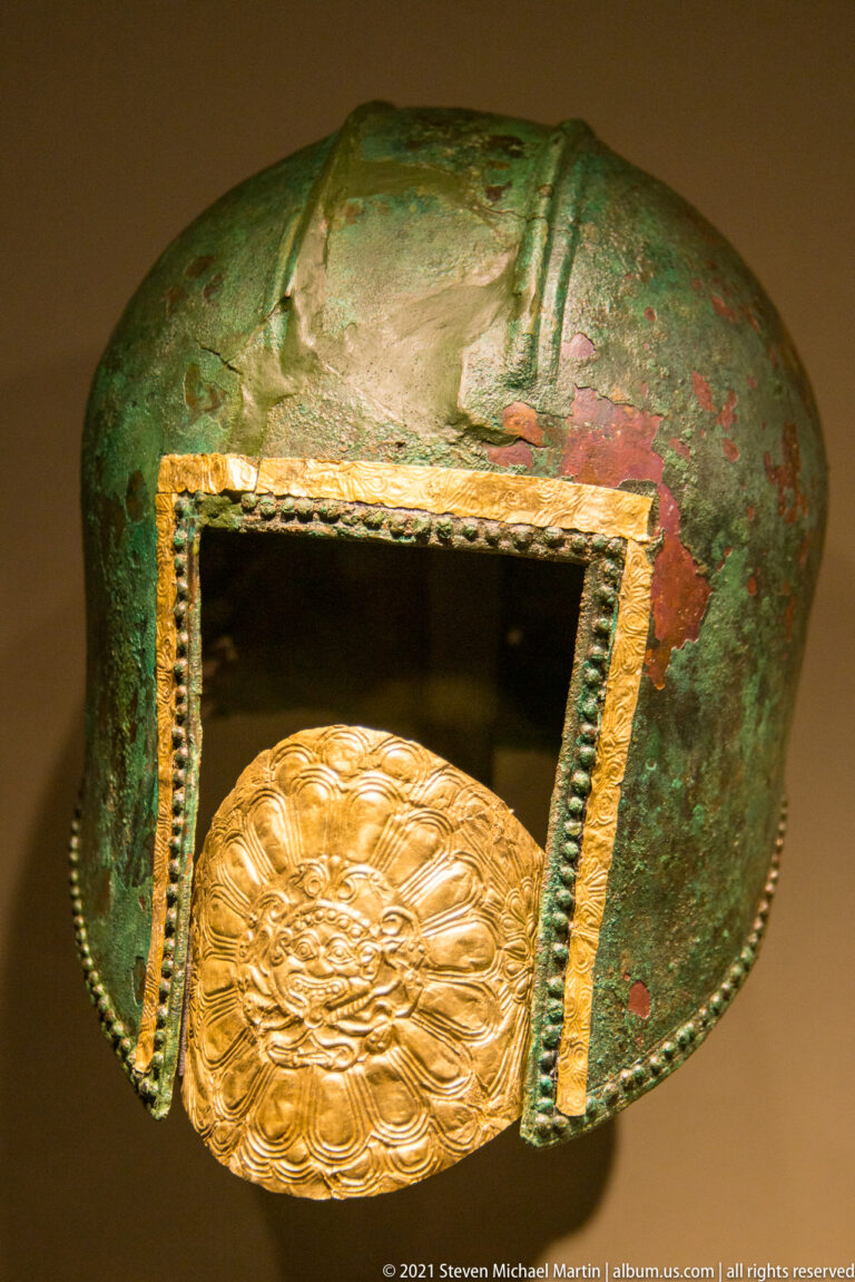 Green-patinated bronze fitting with a rectangular opening, beaded trim, red traces, and an inset oval gold medallion.