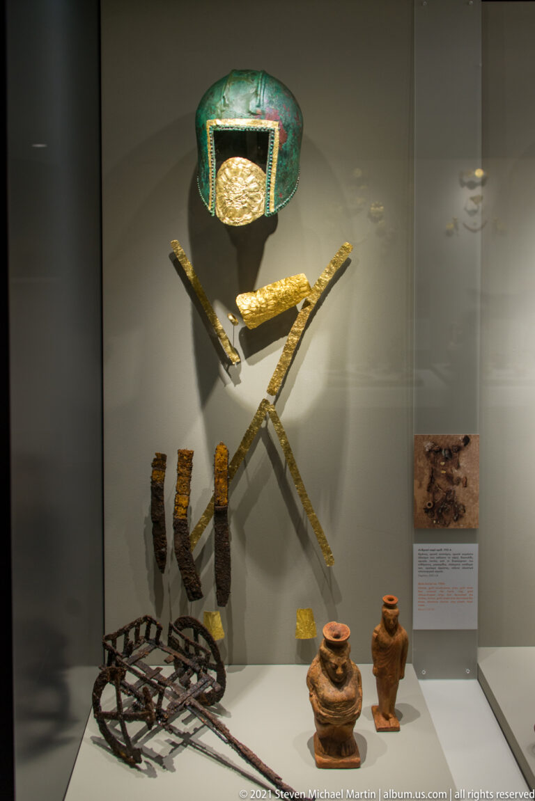 Bronze-and-gold fitting above crossed gold bands, iron objects, and small ceramic vessels in a museum case.