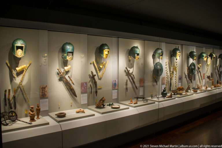 Long museum case with multiple vertically arranged funerary assemblages of jewelry, weapons, vessels, and metal fittings.