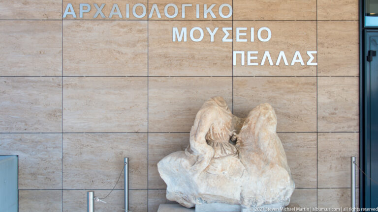 Weathered pale stone sculpture displayed beneath large Greek lettering for the Archaeological Museum of Pella.
