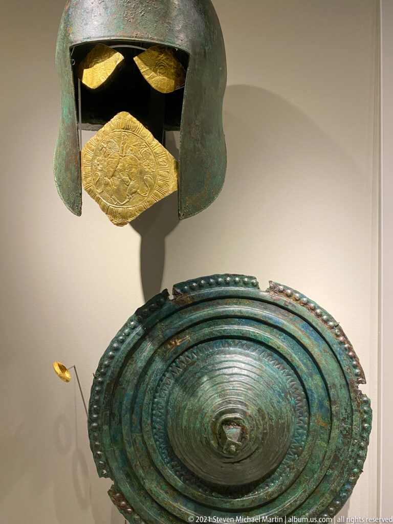 Green bronze helmet above a large circular bronze shield with concentric rings and central boss, accompanied by gold fittings.