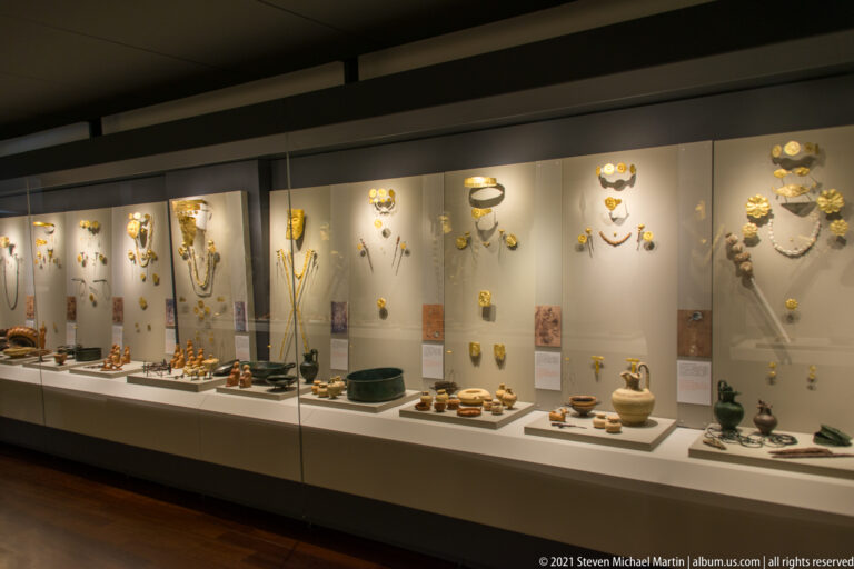 Wide museum case filled with vertically grouped gold jewelry, necklaces, vessels, and other archaeological grave goods.