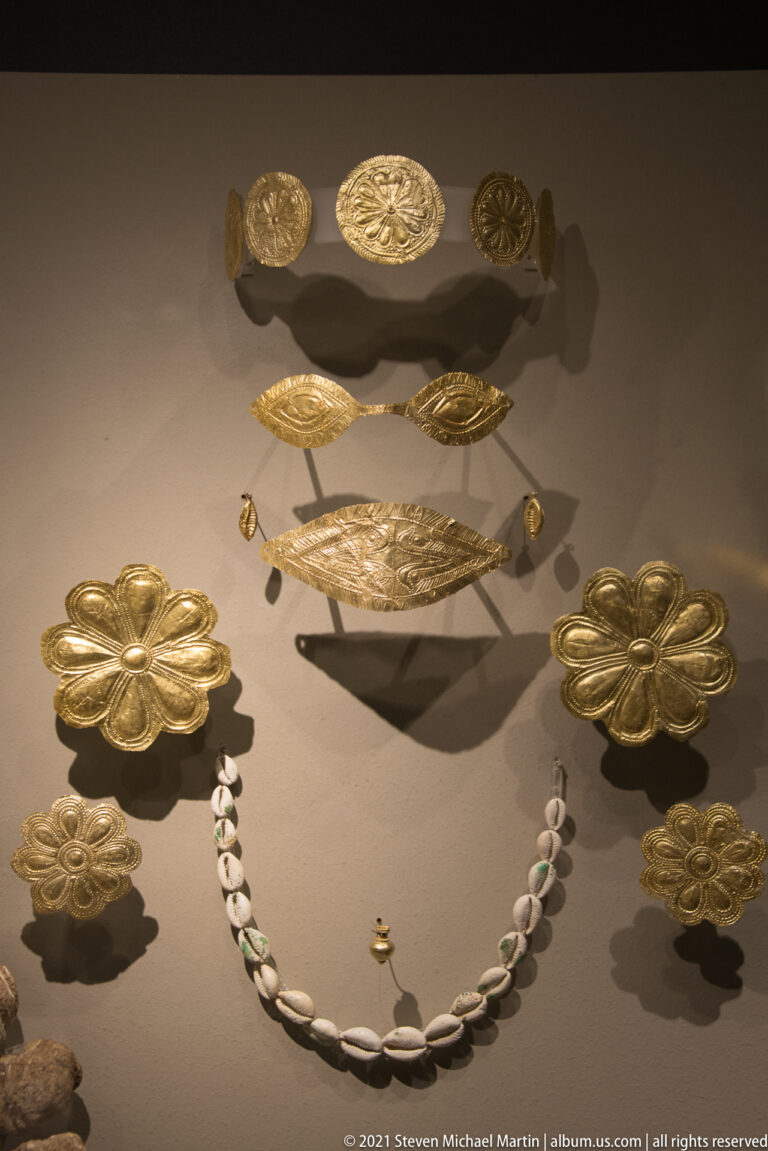 Gold rosettes and narrow embossed bands displayed above a necklace of pale elongated beads.