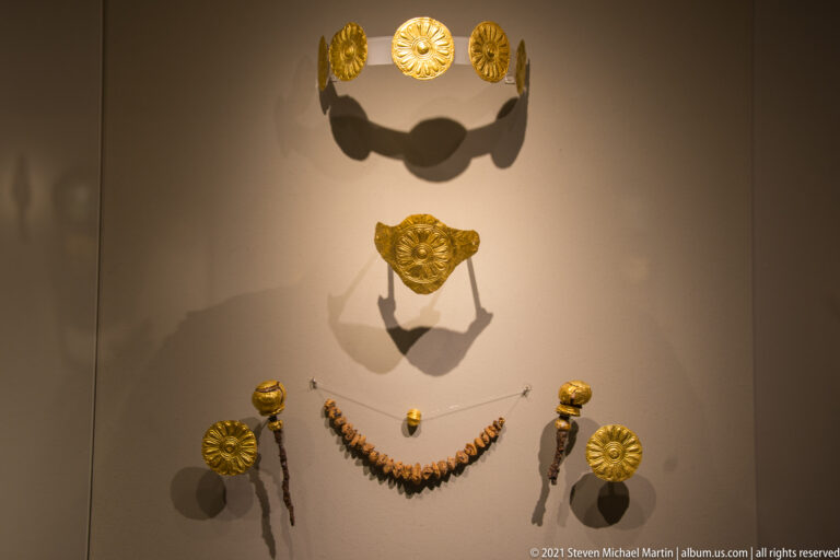 Curved row of gold rosettes above a central gold plaque, bead necklace, and smaller circular ornaments.