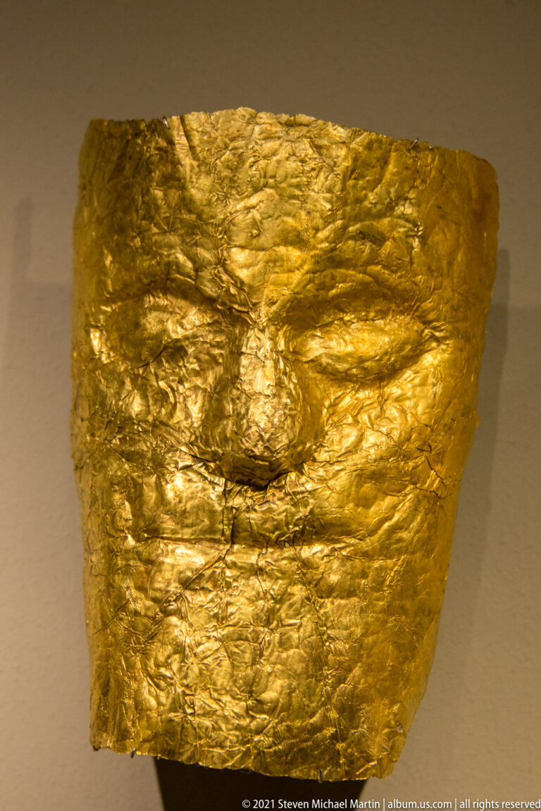 Hammered gold sheet shaped into a stylized human face with modeled eyes, nose, mouth, and brows.