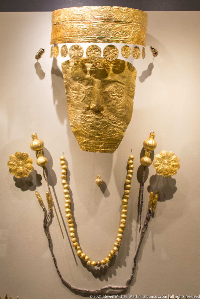 Gold face covering displayed above a curved gold necklace, floral rosettes, and paired hanging ornaments.