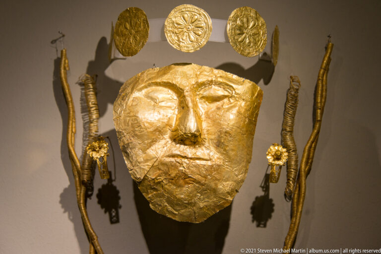 Large modeled gold face covering centered between long narrow ornaments and embossed circular gold disks.